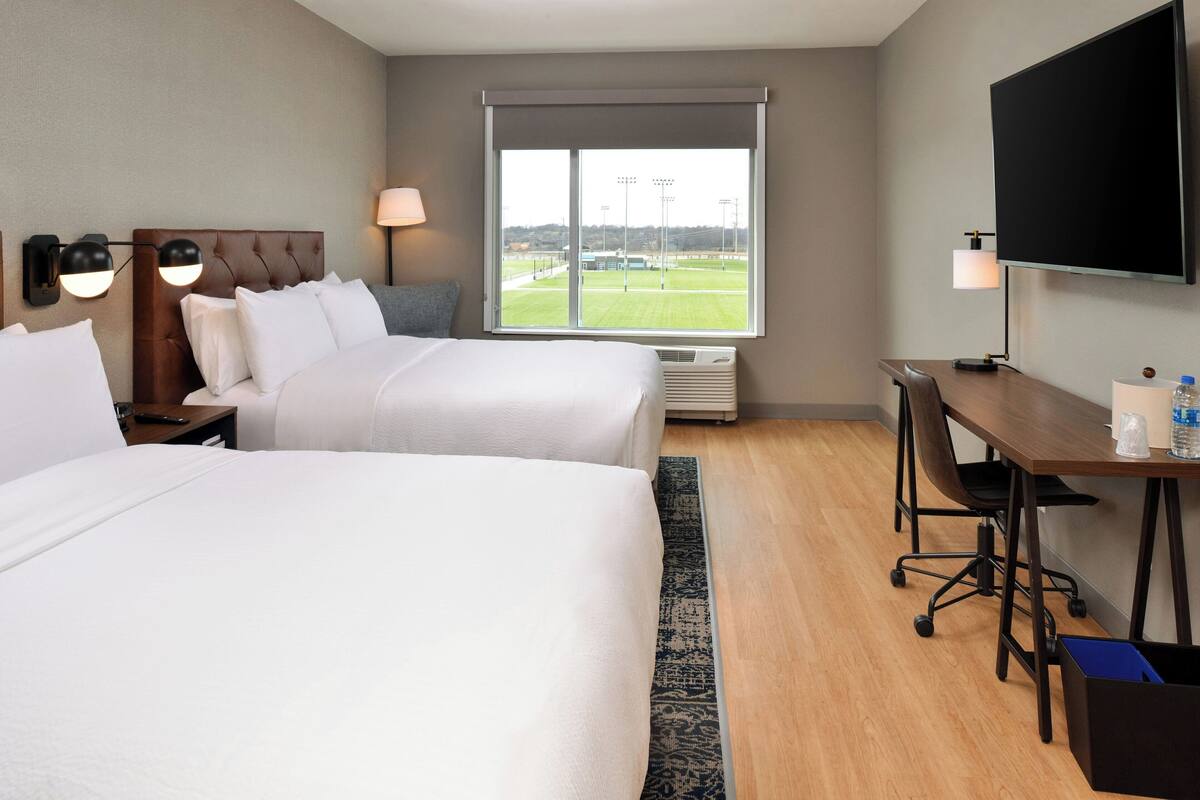 A hotel room with two beds, a desk, a TV, and a window with a view of a field.