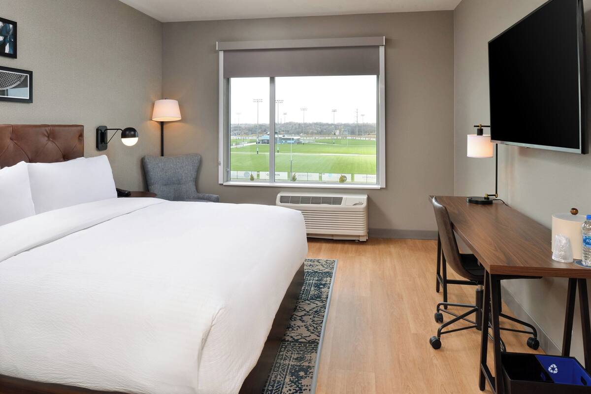 A hotel room with a large bed, a desk with a computer, a TV, and a window with a view of a sports field.
