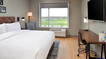 Habitación de hotel con una cama grande, un escritorio con computadora, un televisor y una ventana que da a un campo deportivo.