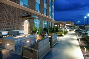 Un patio al aire libre con mobiliario y una chimenea, junto a un edificio con grandes ventanales y fachada de ladrillo.