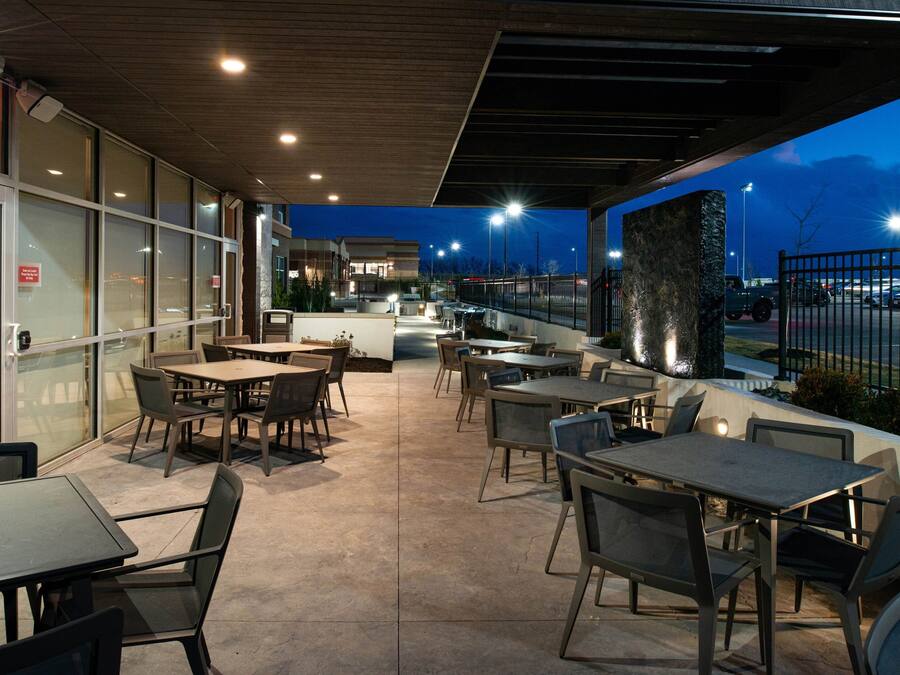 Uma área de pátio externa com mesas, cadeiras, um bar e vista para o estacionamento.