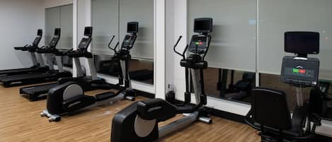 A row of stationary bikes in a gym with wooden flooring and large windows.