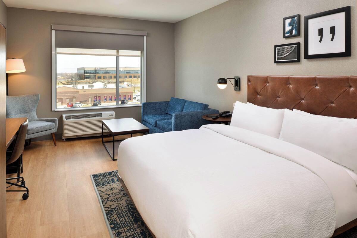 A hotel room with a large bed, a desk, a chair, a sofa, a window with a view of buildings, and framed pictures on the wall.