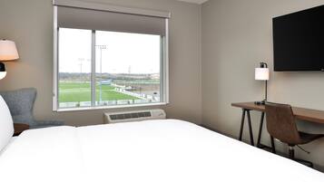 A hotel room with a large bed, a desk, a chair, a TV, and a window with a view of a sports field.