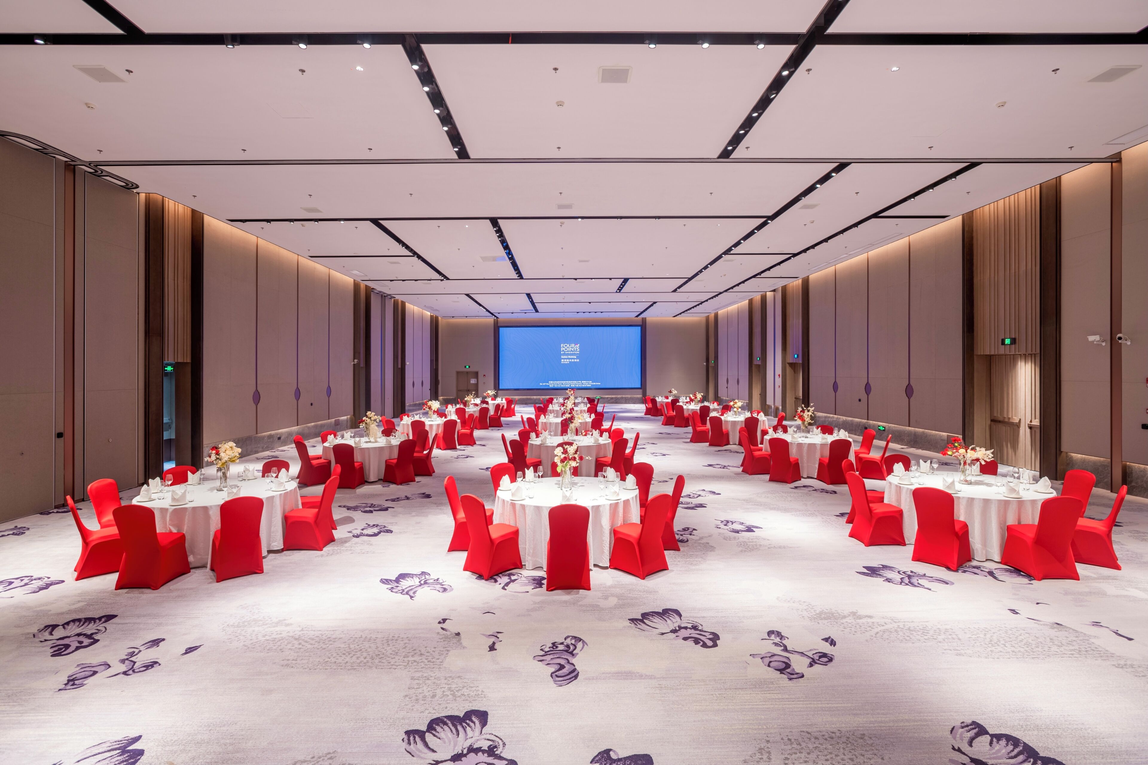A conference room with multiple tables, red chairs, and a large screen at the front.