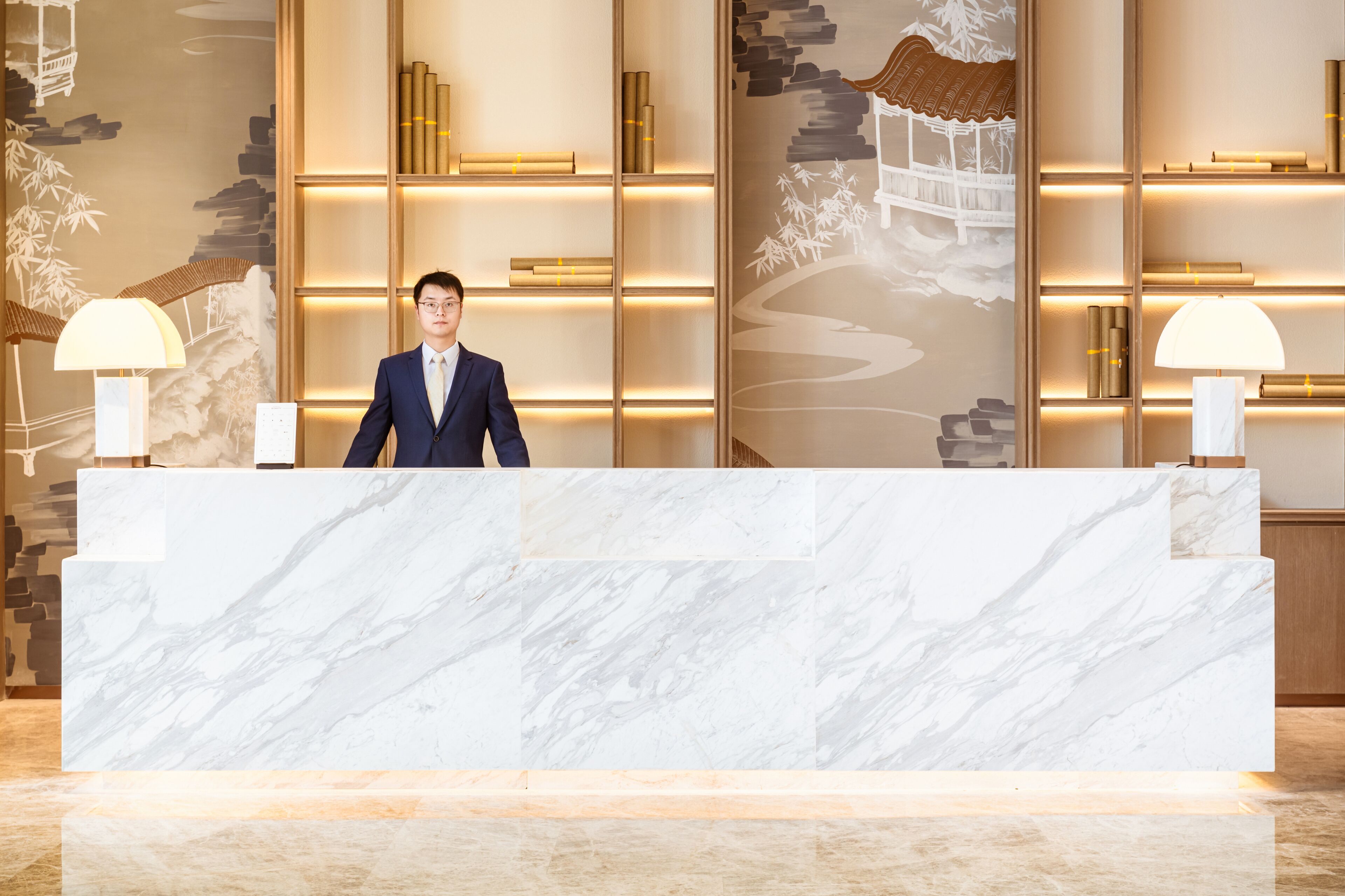 A reception desk with marble top, surrounded by shelves with books and decorative items, and two wall-mounted lamps.