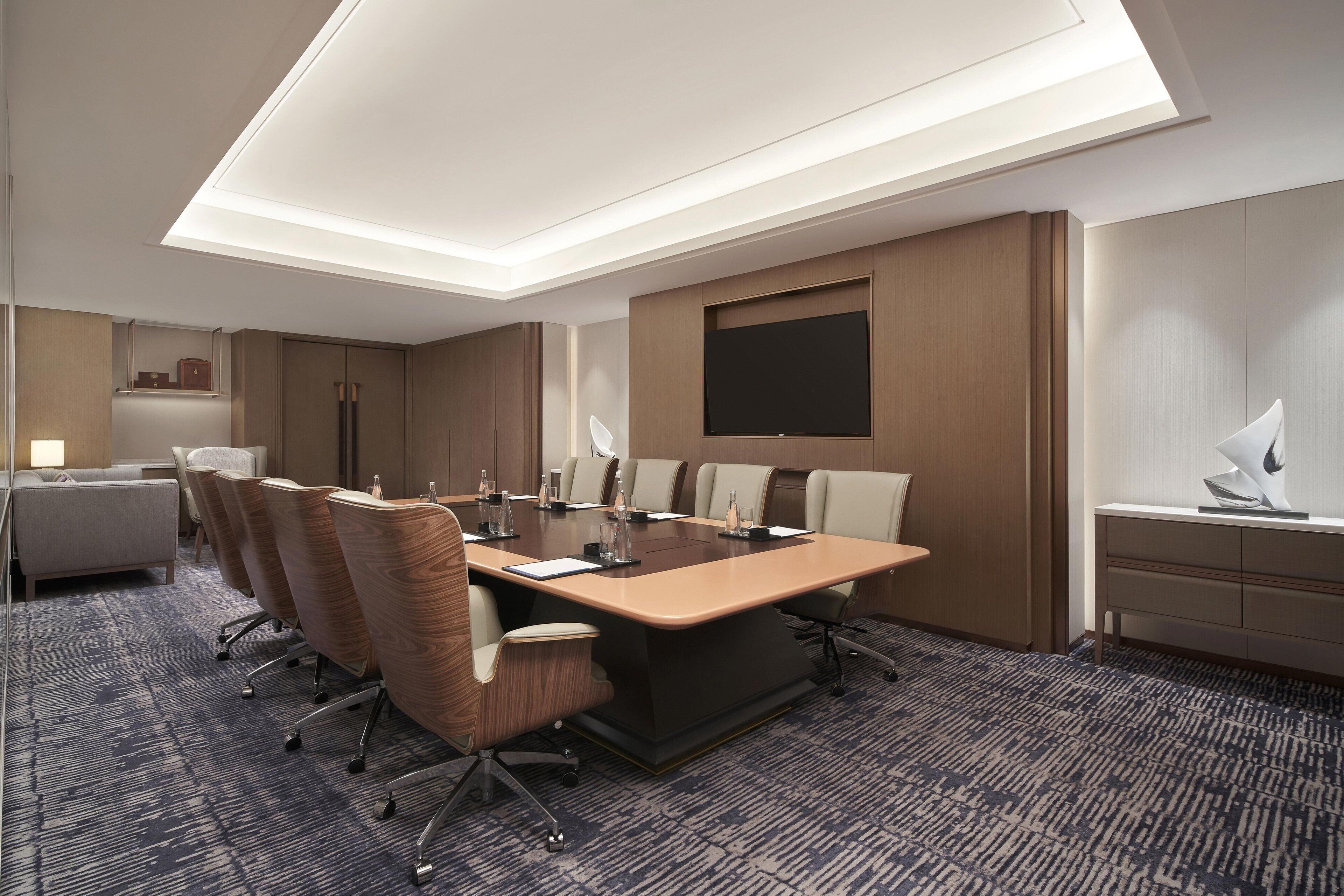 A conference room with a long table, six chairs, a large flat-screen TV, and a desk with a lamp.