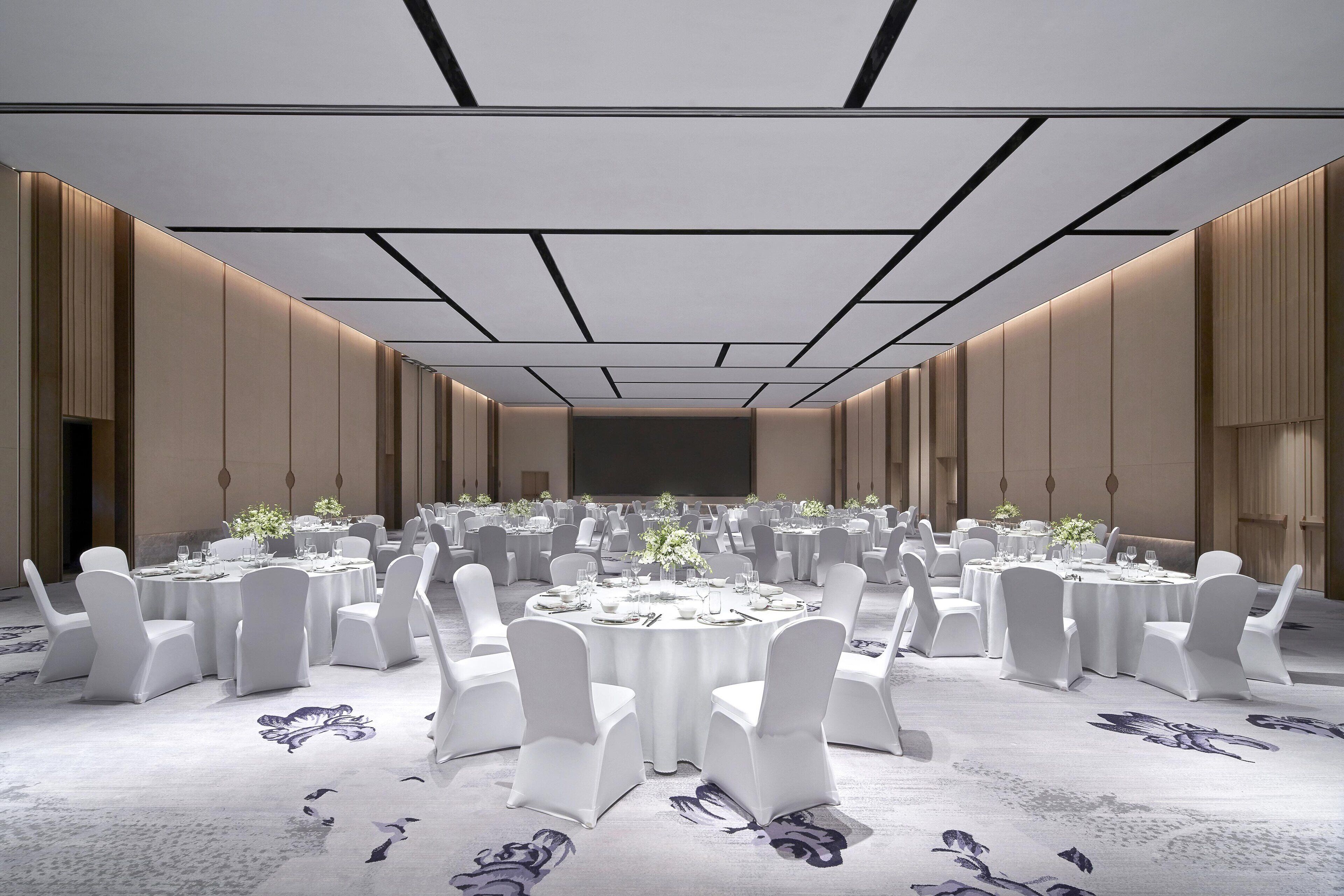A spacious banquet hall with round tables and white chairs, a large screen at the front, and a carpeted floor.