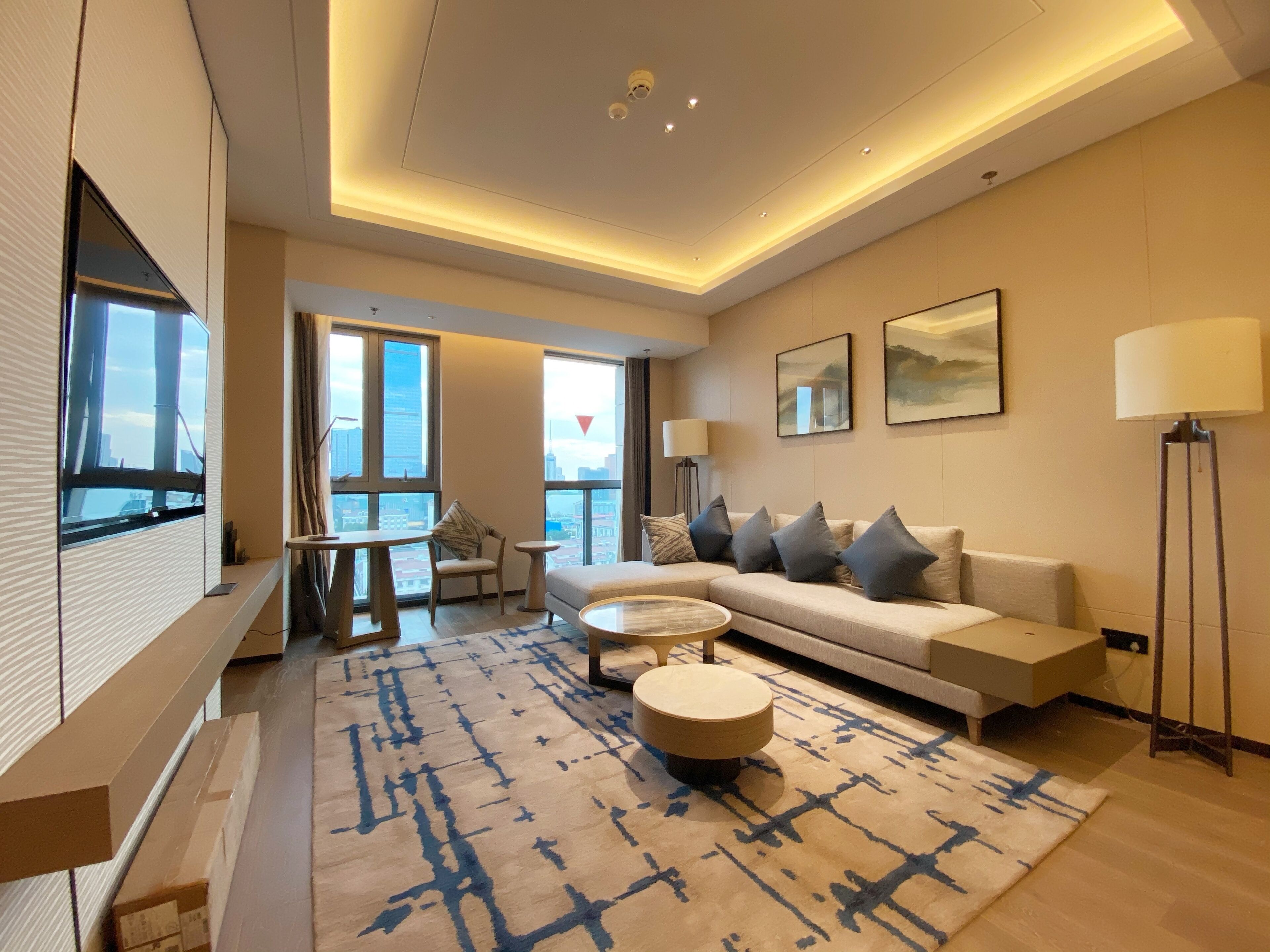 A modern living room with a sofa, coffee table, and a patterned rug. There are large windows offering a view of the city.