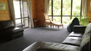 A cozy living room with a leather sofa, a wicker chair, and a small table. Large windows allow natural light and views of greenery outside. - Duvauchelle Dream (Duvauchelle)