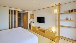 A hotel room with a large bed, a TV mounted on the wall, a bedside table with a lamp, and a wooden shelf with decorative items.