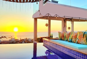 Ein Infinity-Pool mit Blick auf den Sonnenuntergang über dem Meer, eine Sitzecke mit bunten Kissen und eine Bar mit Flaschen.