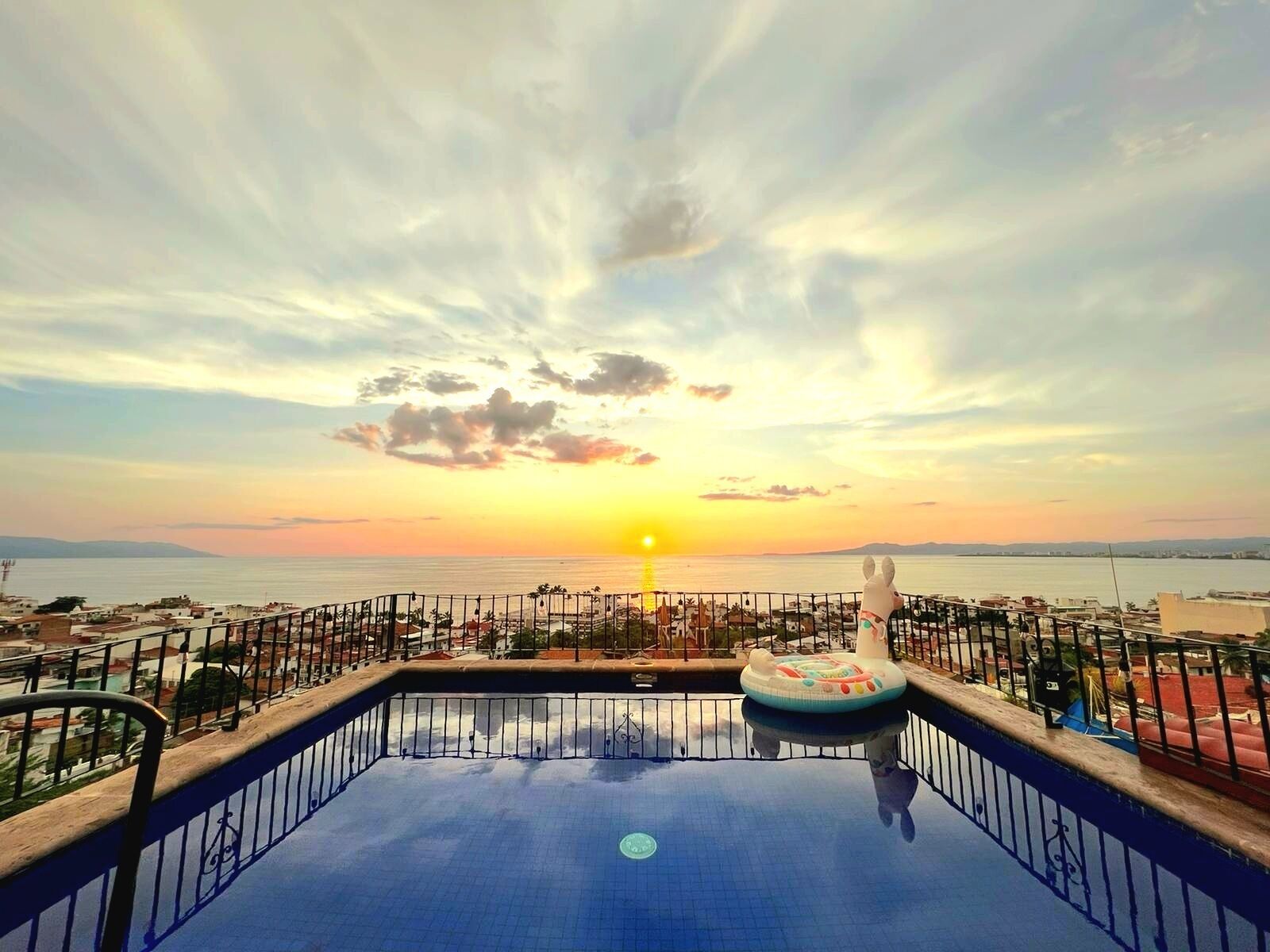 Ein Pool auf dem Dach mit Blick auf den Sonnenuntergang über dem Meer, umgeben von einem Geländer und einem Rettungsschwimmerstuhl.