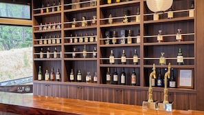 Zona de bar con estantes llenos de botellas de licor, una barra de madera y vistas a un bosque por la ventana.