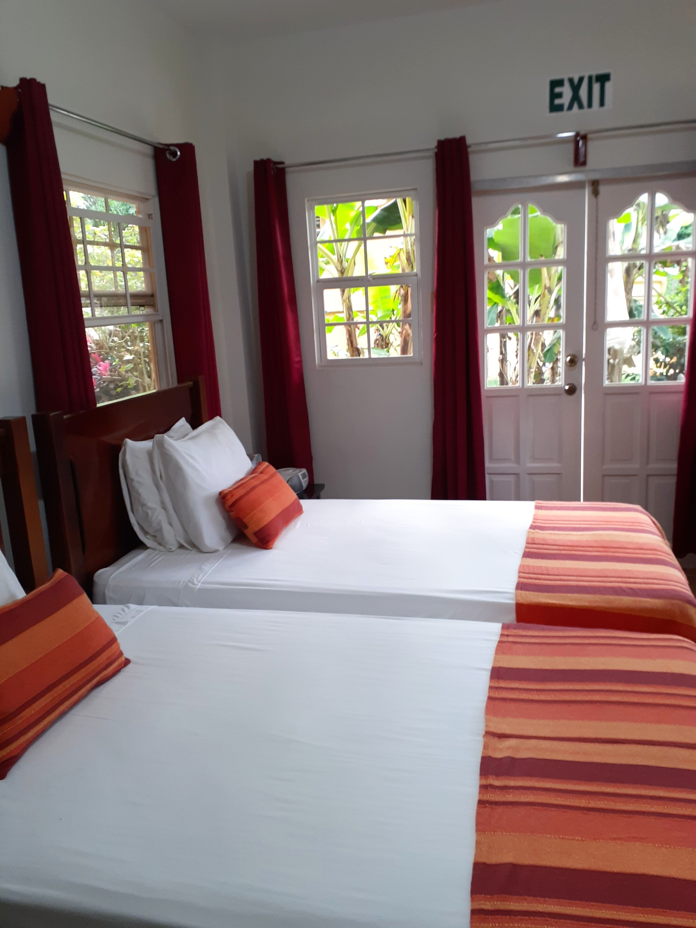 Ein Hotelzimmer mit zwei Betten, roten Vorhängen und Blick auf Grün hinter den Fenstern.