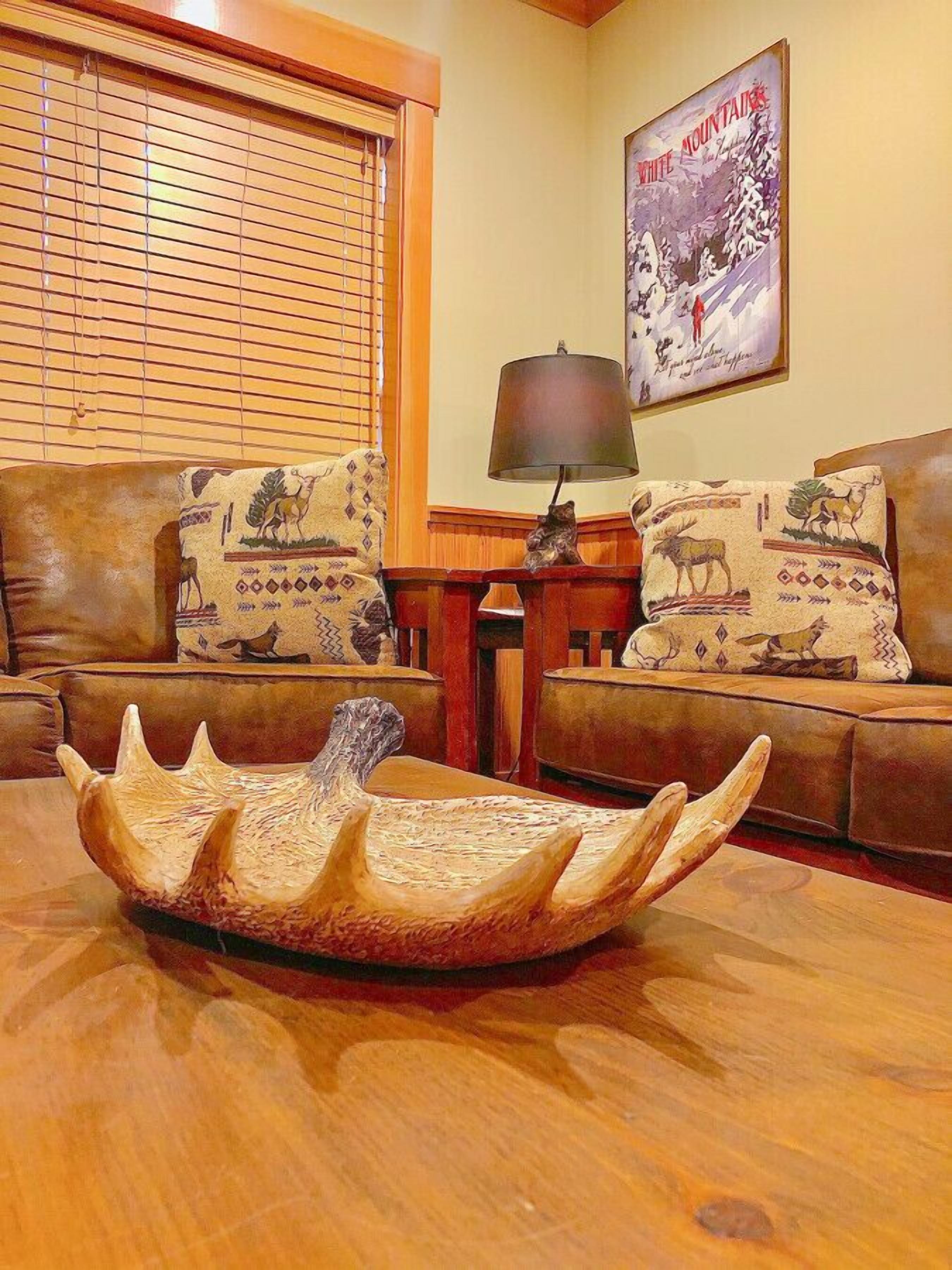 A living room with two brown leather sofas, a wooden coffee table with a carved animal head centerpiece, a lamp, and a framed poster on the wall.