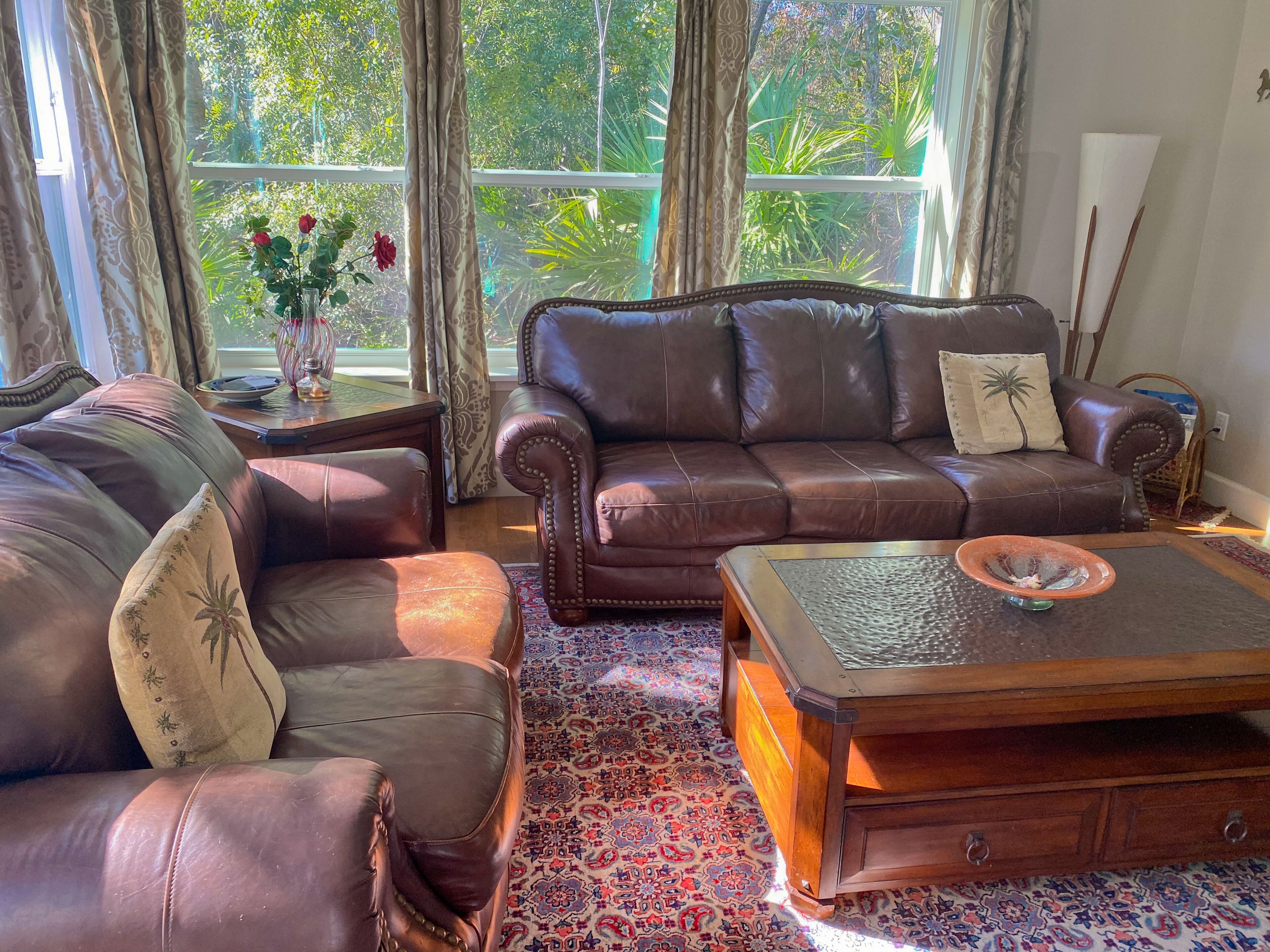 A living room with a brown leather sofa, a matching armchair, and a wooden coffee table. There is a patterned rug on the floor, and a window with curtains and a vase of flowers.