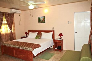 Un dormitorio con cama de madera, dos mesitas de noche con lámparas, ventilador de techo, equipo de aire acondicionado, una puerta y un tapete verde.