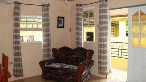 Un salon avec un canapé à motifs, un ventilateur de plafond et une vue sur l’extérieur par une fenêtre ornée de rideaux.