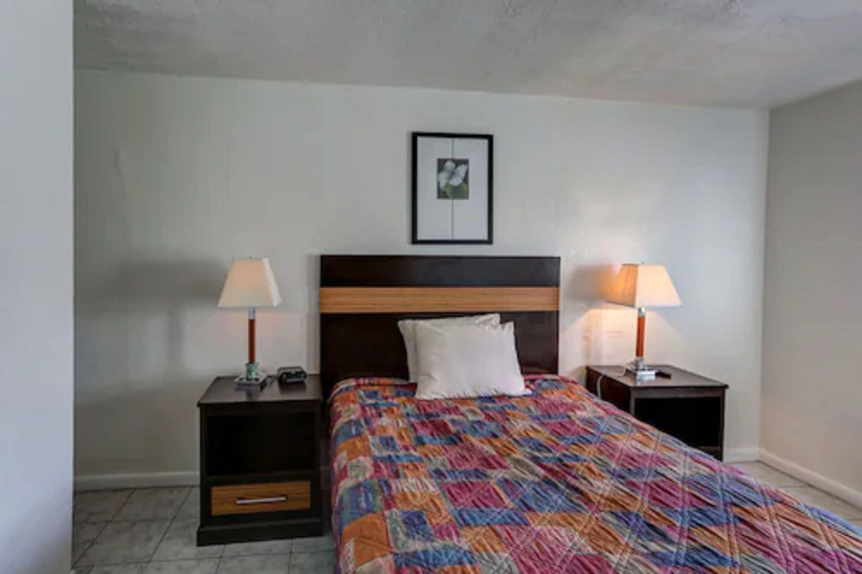 A bedroom with a wooden headboard, a colorful quilt, two bedside tables with lamps, and a framed picture on the wall.