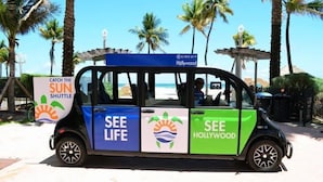 Un autobús lanzadera colorido con la leyenda "SEE LIFE" y "SEE HOLLYWOOD" en sus laterales, estacionado en una playa soleada con palmeras.