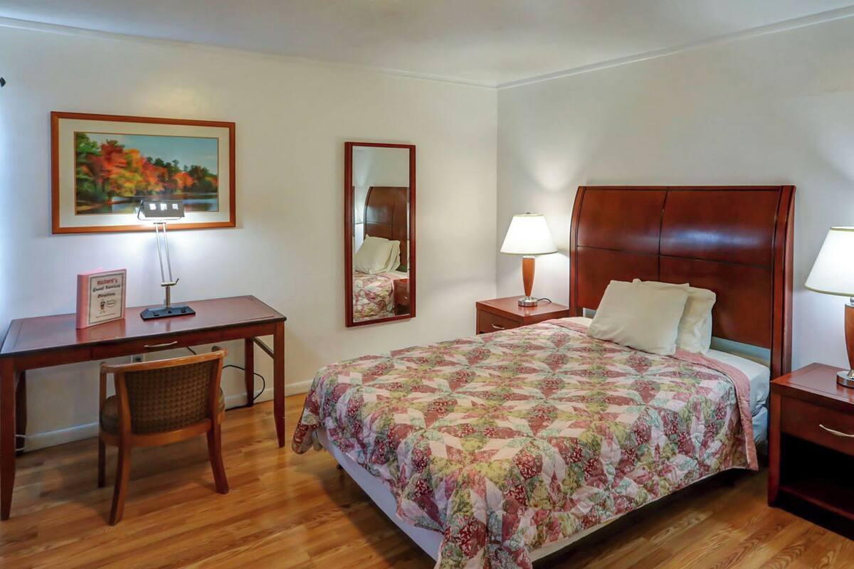 A hotel room with a wooden bed, two bedside tables with lamps, a desk with a chair, a mirror, and a framed picture on the wall.
