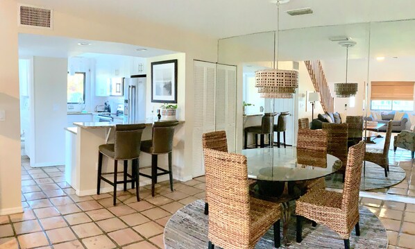 Eine moderne Küche mit weißen Schränken, eine Essecke mit Korbstühlen und ein Wohnzimmer mit Sofa und Couchtisch.