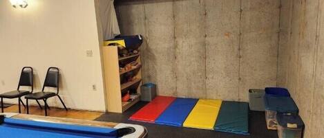 A room with a pool table, chairs, a shelf with storage boxes, and a padded floor area.