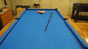 Uma mesa de bilhar com pano azul, um taco e um suporte colorido para as bolas.