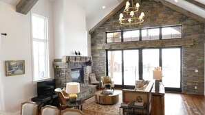 A spacious living room with a stone fireplace, a large window, and a chandelier. - New! Luxury Mountain Escape in Majestic Playground (Tetonia)