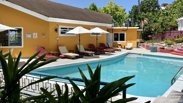 A pool area with lounge chairs and umbrellas, surrounded by greenery and a yellow building. - Palm Garden (Montego Bay)