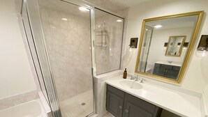 Un baño moderno con ducha envuelta en vidrio, un lavamanos blanco y un espejo con marco dorado, además de un mueble de color gris oscuro.