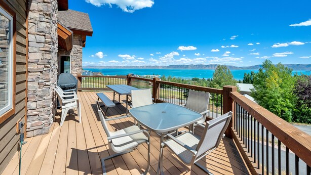 A wooden deck with a stone wall, outdoor seating, and a view of a lake and mountains.