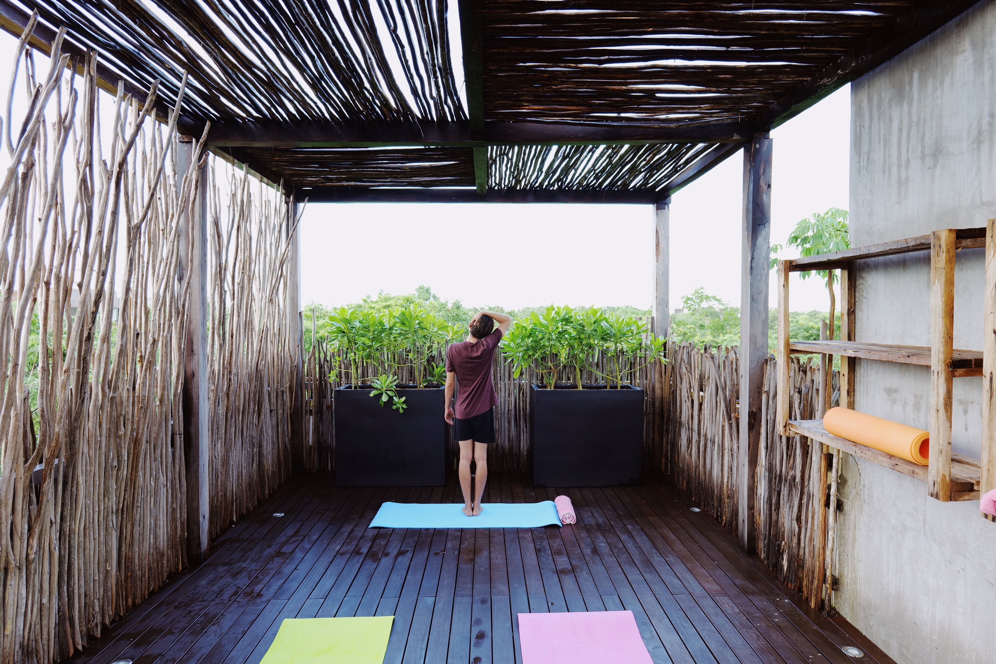 Un espacio al aire libre techado con cubierta de madera, una persona de pie sobre una colchoneta de yoga y equipo de ejercicio.