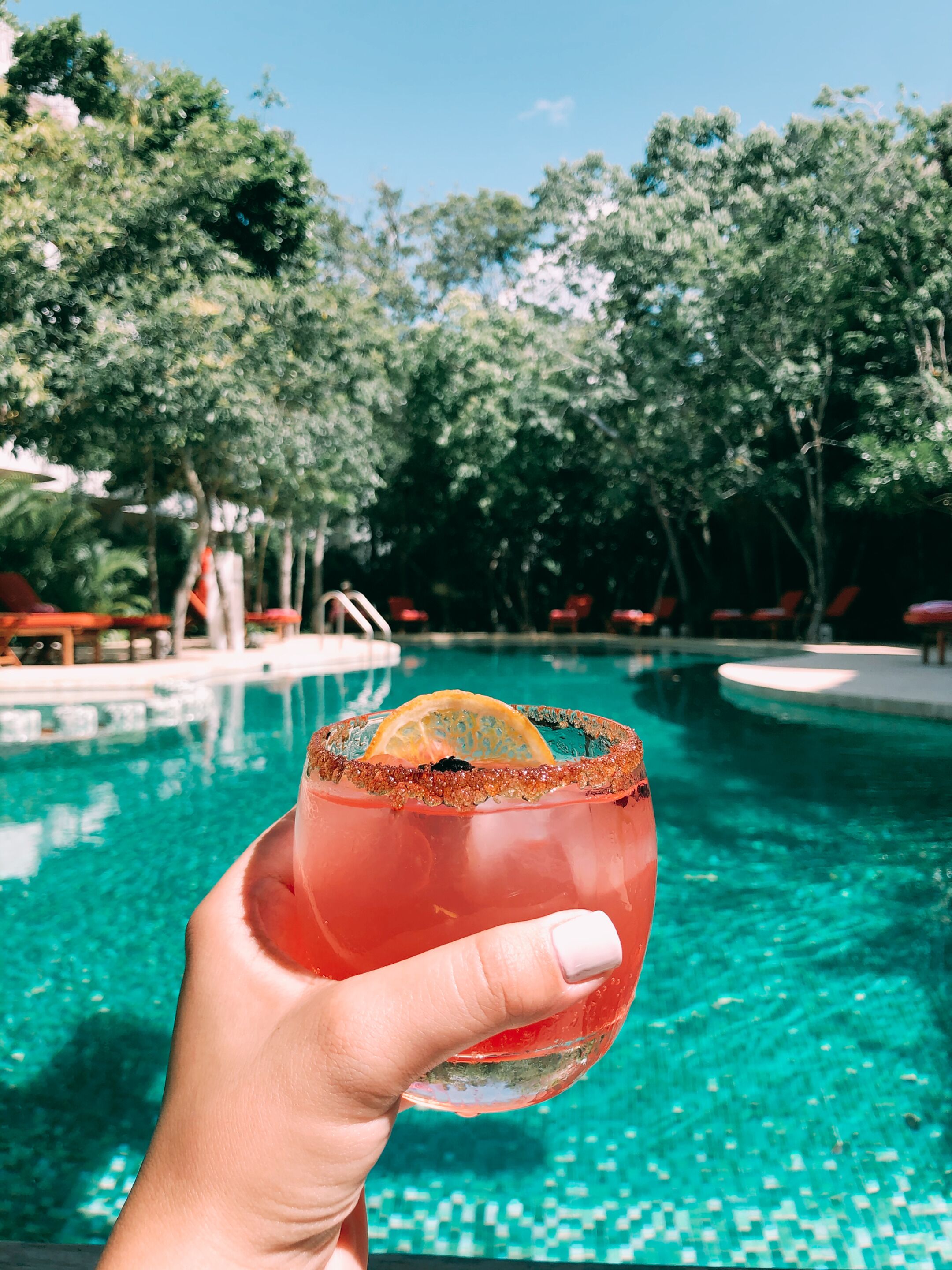Una mano sostiene un cóctel con borde de sal y una rodaja de naranja, frente a una piscina con agua azul cristalina y sillas rojas alrededor.