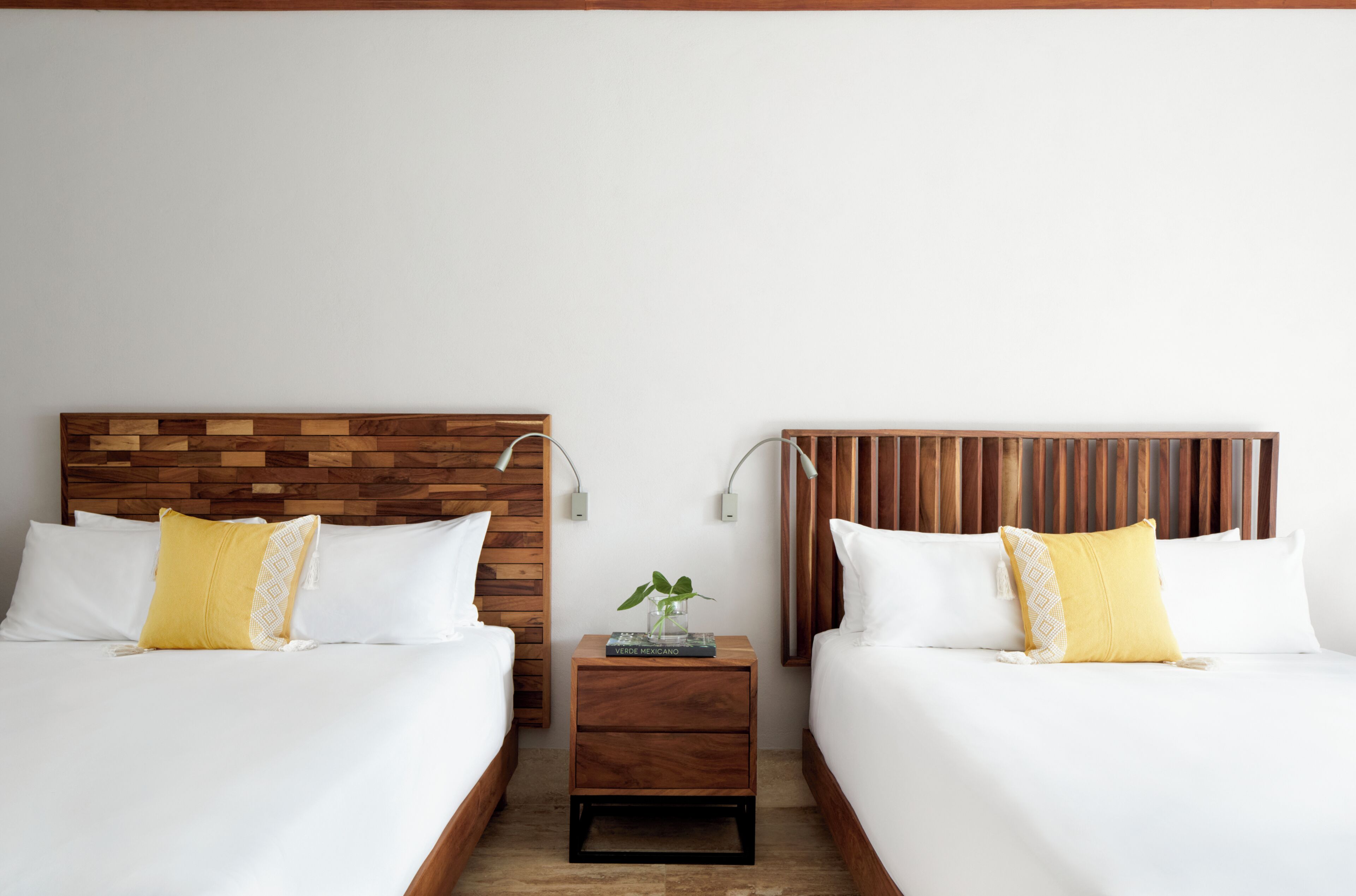 A hotel room with two beds, wooden headboards, white bedding, and a small bedside table with a book and a plant.