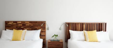 A hotel room with two beds, wooden headboards, white bedding, and a small bedside table with a book and a plant.