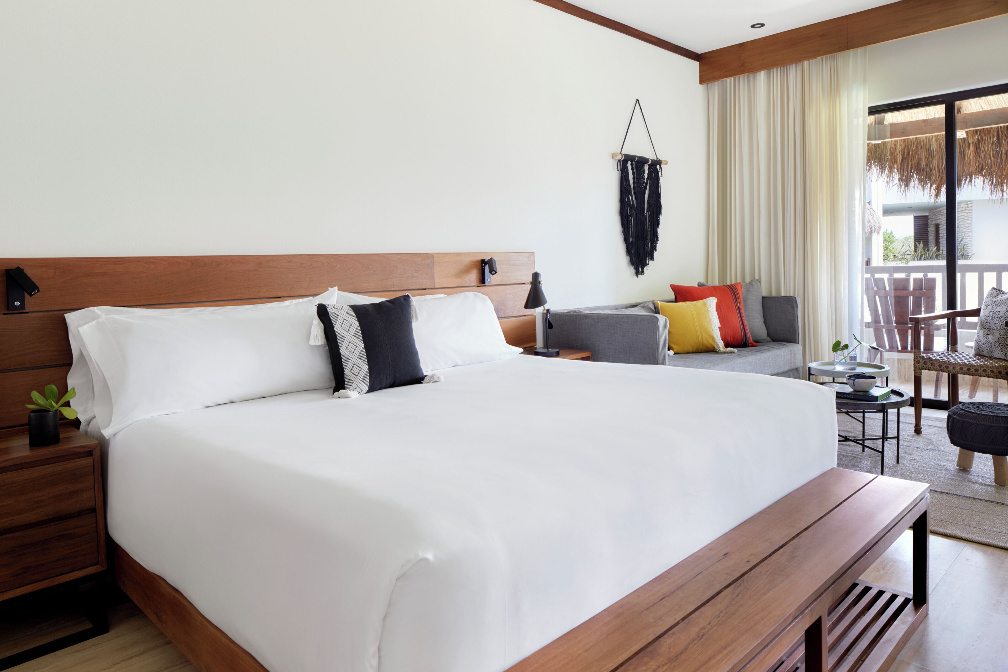 A hotel room with a large bed, a wooden headboard, a nightstand with a plant, a small table, and a balcony with a chair.