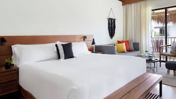 Habitación de hotel con una cama grande, cabecera de madera, mesita de noche con una planta, una pequeña mesa y un balcón con una silla.