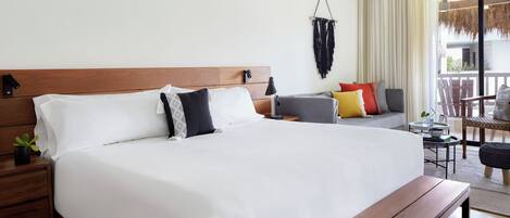A hotel room with a large bed, a wooden headboard, a nightstand with a plant, a small table, and a balcony with a chair.