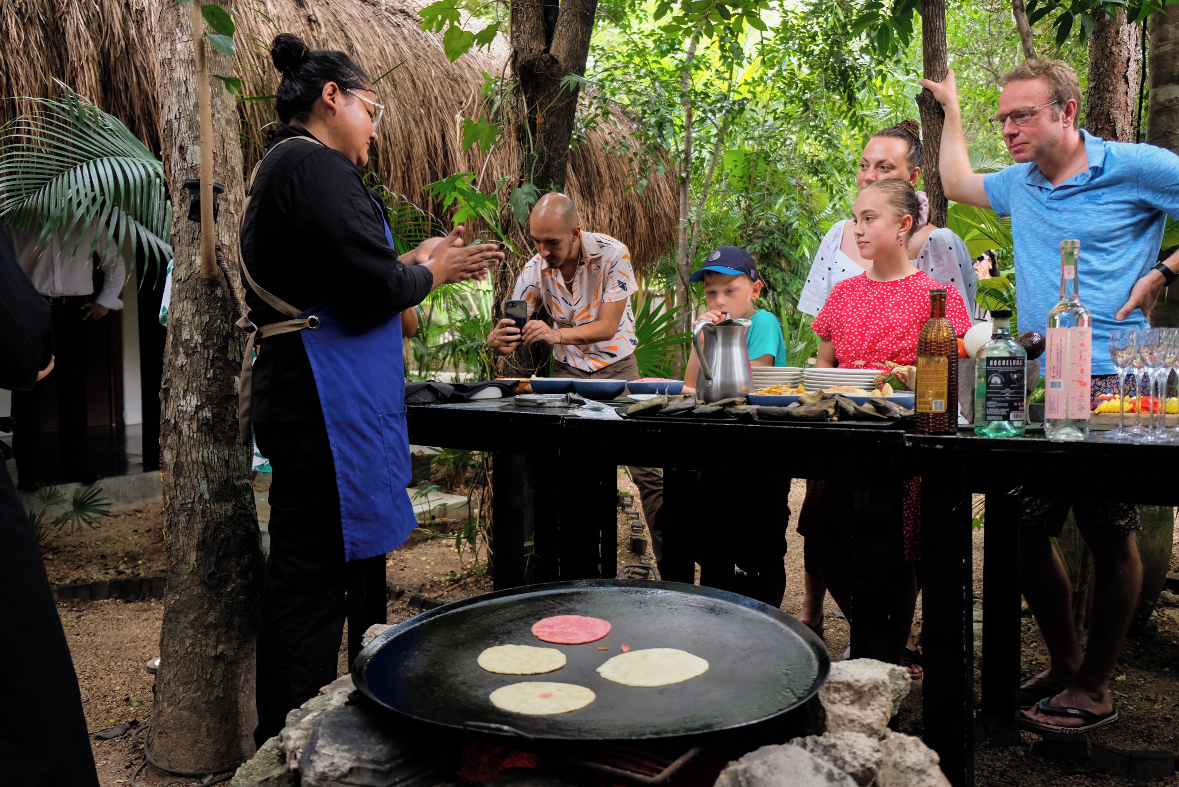 A group of people around a grill with flatbreads cooking, a table with condiments, and a thatched-roof structure in the background.