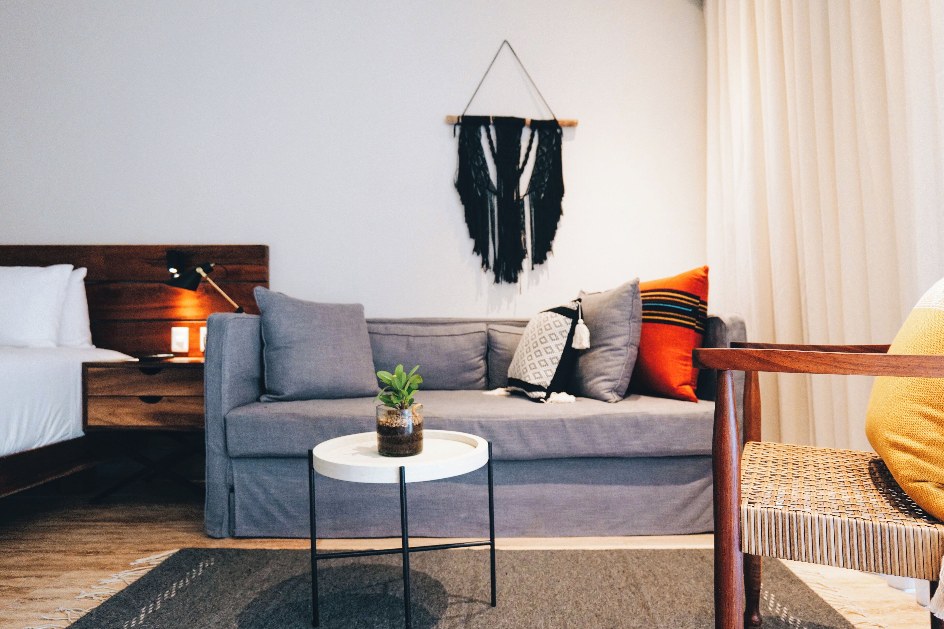 A modern hotel room with a grey sofa, a small round table with a plant, a hanging macrame wall decoration, and a wooden nightstand with a lamp.