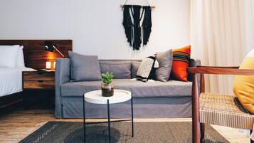A modern hotel room with a grey sofa, a small round table with a plant, a hanging macrame wall decoration, and a wooden nightstand with a lamp.