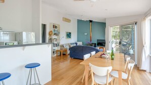 Una sala de estar moderna con sofá, una mesa de comedor con sillas, una zona de cocina con refrigerador y un amplio ventanal con vista a zonas verdes.