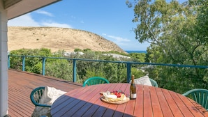 Una terraza de madera con una mesa preparada para una comida, con vistas a un paisaje montañoso y el mar.