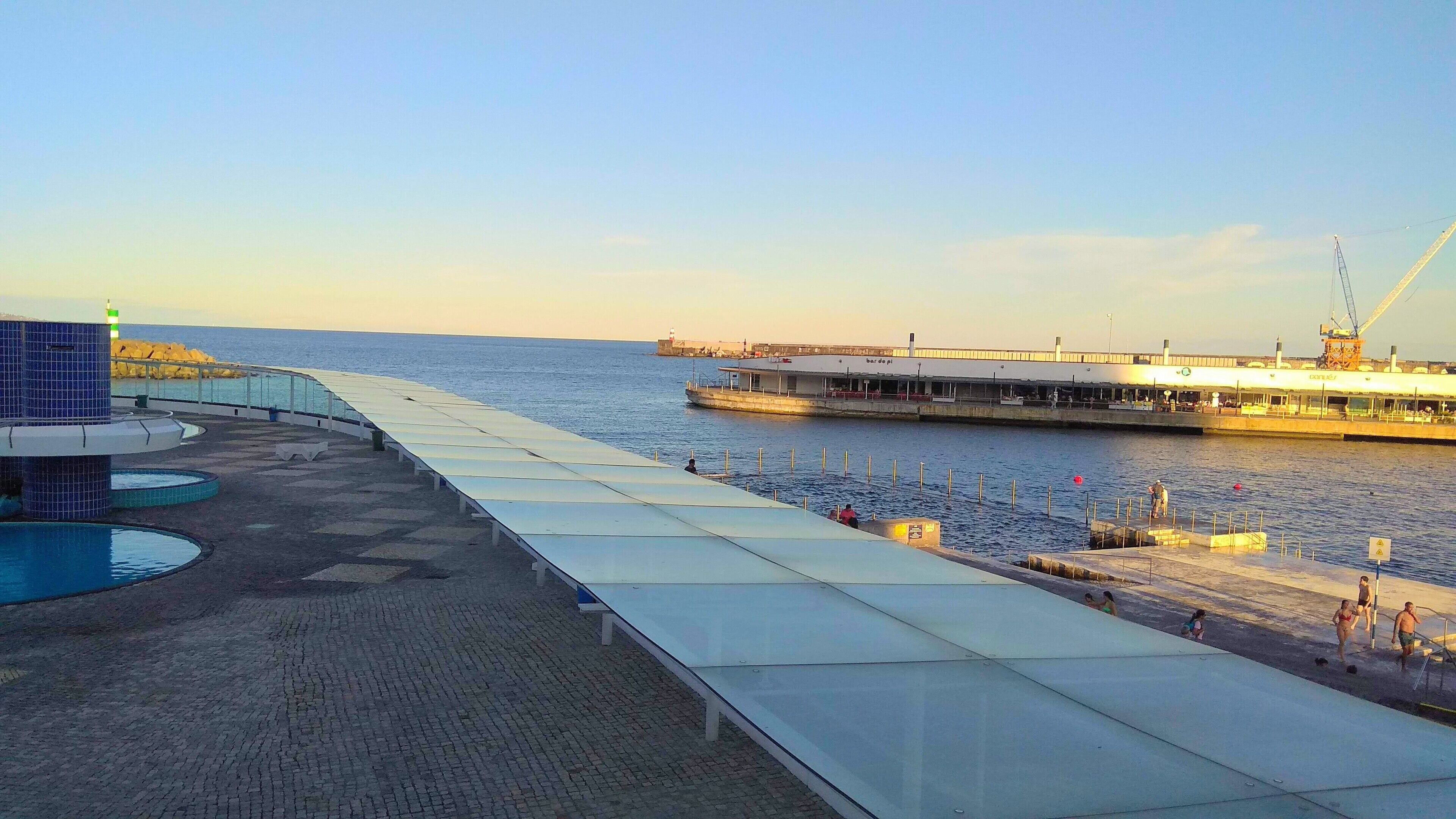 A waterfront promenade with a covered walkway, a pool, and a pier with a boat.
