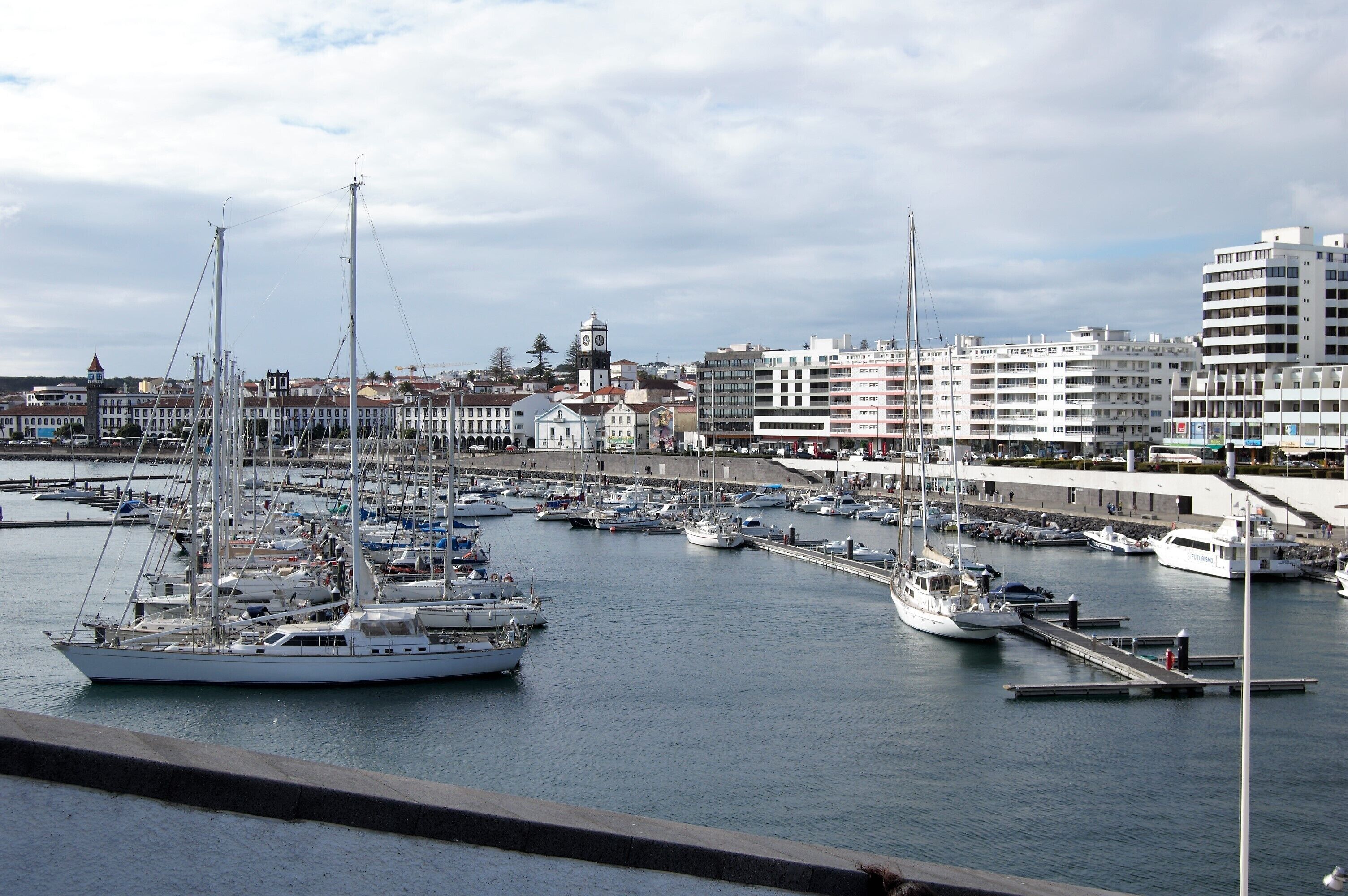 A marina with multiple boats docked, a prominent clock tower, and modern buildings in the background.