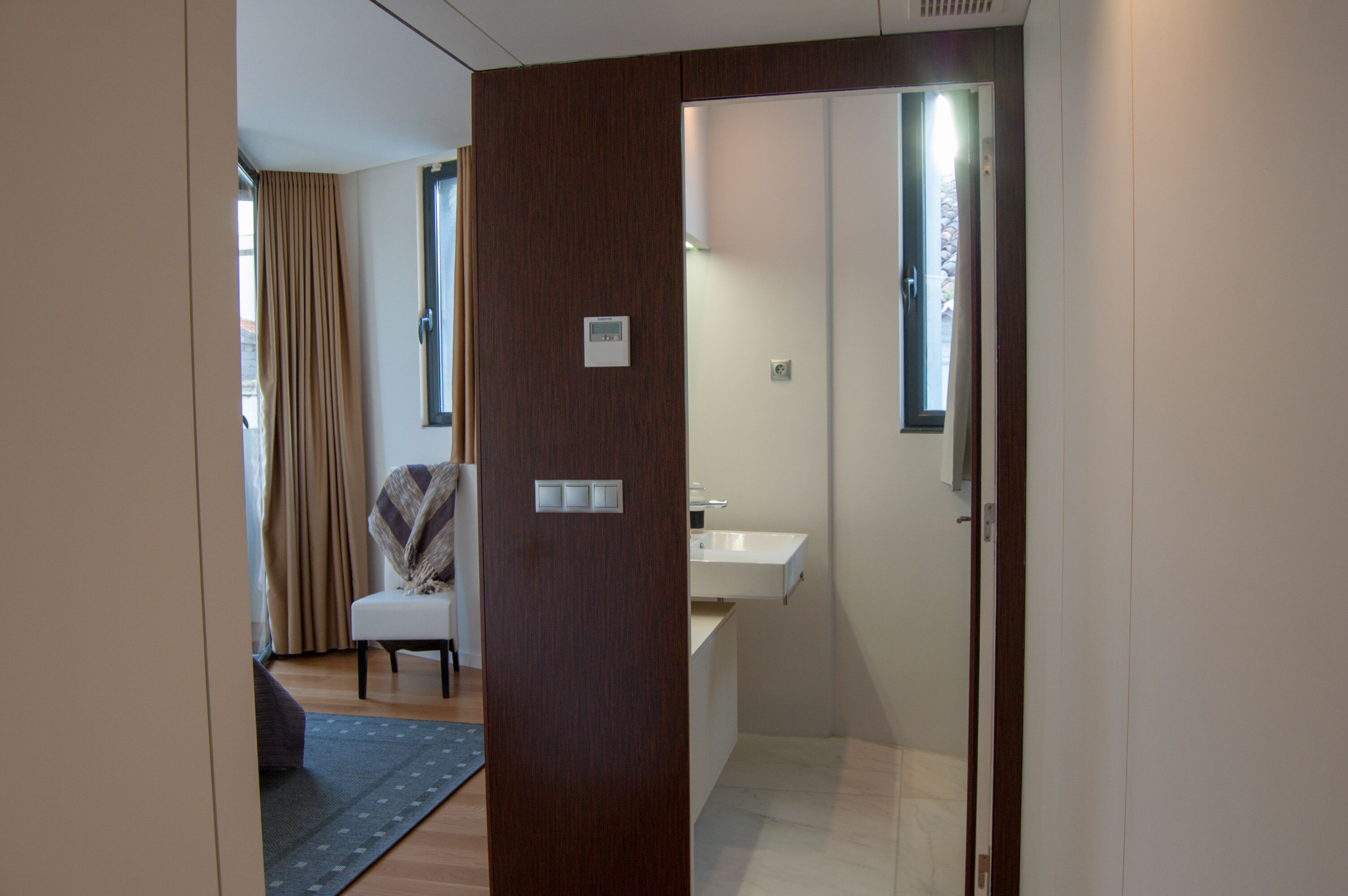 Una habitación de hotel con puerta de madera, una ventana pequeña, un lavamanos blanco y una silla con una manta.