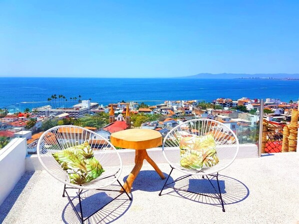 Une terrasse avec deux chaises blanches à armature métallique et une petite table ronde, d’où l’on peut admirer une ville côtière et la mer.