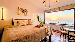 Quarto com cama grande, mesa de cabeceira, luminária na parede, objeto decorativo na parede, e vista para o oceano ao pôr do sol.